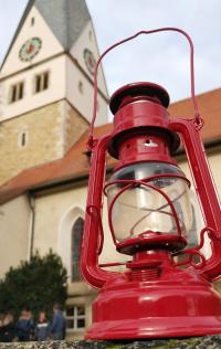 Auf einer Mauer steht eine brennende Öllampe. Im Hintregrund sieht man die Konturen des KIrchturms.