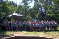 Eine Gruppe von über 80 Pfadfindern steht bei Sonnenschein im Wald. Im hintergrund sieht man eine "fliegendes" Luftschiff aus schwarzen Zeltplanen.